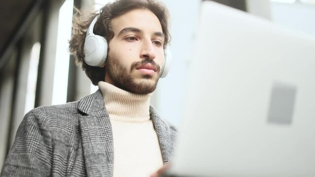 Handsome young arabian businessman with headphones working on laptop sitting near modern business centre Busy focused muslim man have distance remote freelance work on computer outdoors alone
