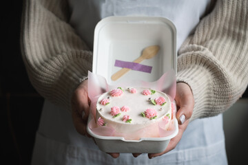 Woman pastry shef wearing white apron and holding small bento cake decorated with cream cheese pink flowers. Woman hands hold birthday cake in a white gift box. Trendy Korean style cake