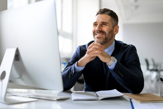 Happy pensive middle aged businessman in suit sitting in front of computer, thinking and smiling, working in office