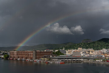 Fototapeta na wymiar Port Louis, Mauritius - March 2023, a rainbow in front of dark grey rainy sky arises from old historic buildings in the harbor of Port Louis 