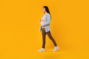 Young pregnant woman in casual wear, walking and touching her belly on yellow studio background, full length, free space