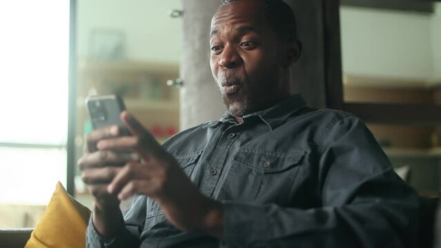 Portrait of mature smiling african american man looking at smartphone cellphone screen with nod of approval sitting on sofa at home and giving thumbs up Advise Approve Recommend Choice concept