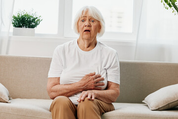 Elderly woman severe pain in the heart sitting on the couch, health problems in old age, poor quality life. Grandmother with gray hair holding on her chest, heart attack and stroke, pressure problem.