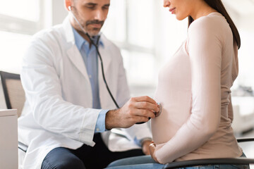 Obraz premium Healthcare and prenatal care concept. Male doctor examining pregnant woman's belly using stethoscope, cropped