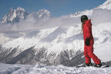 Fototapeta na wymiar Young woman snowboarder wearing red ski suit and helmet riding on a slope against backdrop of stunning mountains in ski resort