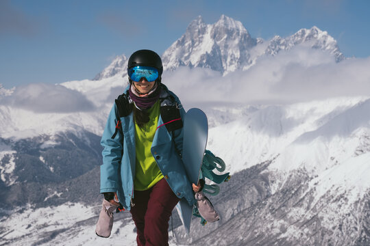 Snowboarder young female smiling and walking with snowboard beautiful mountain peaks covered with snow on background