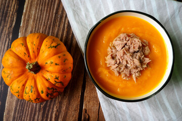 on a brown wooden table is a plate of pumpkin cream soup with tuna next to a small pumpkin.  gluten free, lactose free.  diet .  view from above .  menu calendar