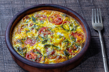 Ceramic bowl with vegetable frittata, simple vegetarian food. Frittata with egg, tomato, pepper, onion, green dill and cheese on wooden table, closeup. Italian egg omelette