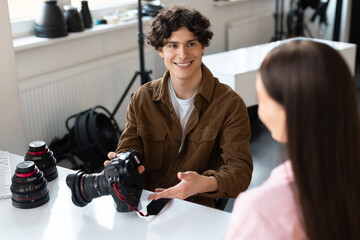 Cheerful male photographer talking with model, discussing result of photoshoot, sitting at desk with camera and lens - Powered by Adobe
