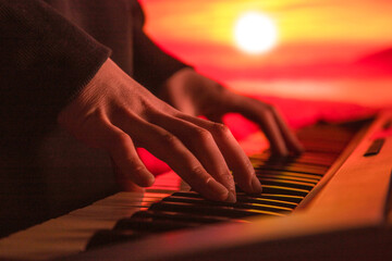 close-up of the hands of a musician playing a synthesizer against the backdrop of a sunset sky - obrazy, fototapety, plakaty