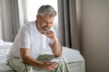 Cheery mature man sitting on bed, using smartphone and smiling