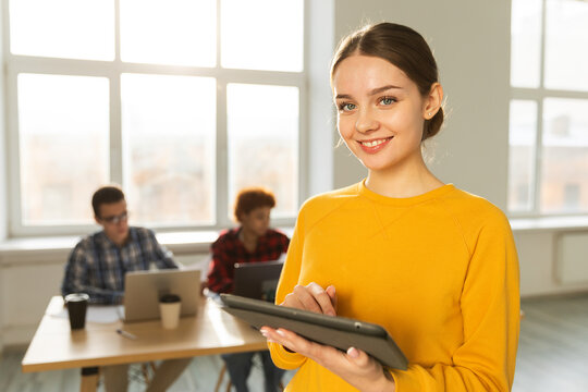 Portrait of female team leader businesswoman standing in office with colleagues meeting in background. Smiling confident business leader woman holding tablet looking at camera standing in office