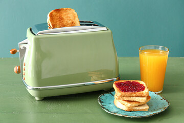 Crispy bread slice in modern toaster, cup of juice and toasts with jam on green wooden table