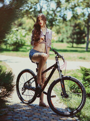 beautiful and young girl with blond hair posing on a bike in the park in summer. In shorts and a shirt.