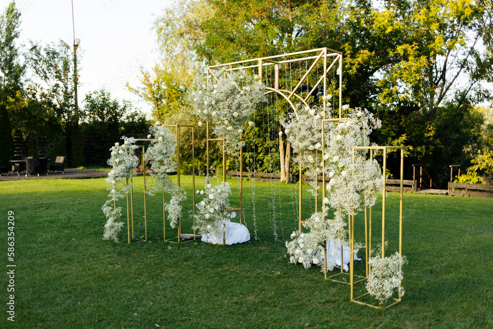 Wall mural Wedding ceremony outdoor. A beautiful and stylish golden wedding arch, decorated by fresh white gypsophila and roses flowers with white chairs, standing in the garden. Celebration day. 