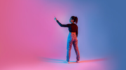Back view of black lady touching imaginary screen, using virtual interface on colorful neon...