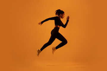 Fototapeta na wymiar Side view of young black sportswoman jumping, posing in mid-air on orange neon background, free space, full length
