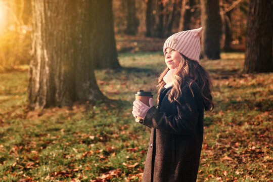Funny little girl child model in coat and hat holding coffee in autumn park, looking away. Positive cute lady kid in fall nature leisure activity. Childhood vitality and lifestyle concept. Copy space