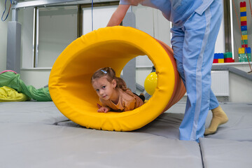 Kid girl rotation in roller tonnel during sensory integration session. child with physiotherapist...