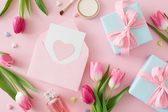 Top view photo of open envelope with postcard gift boxes perfume bottle cosmetic brush pink tulips flowers and colorful hearts on isolated pastel pink background. Mother's Day concept.
