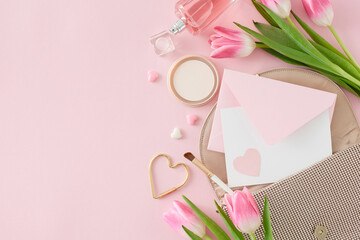 Top view photo of envelope with postcard women's handbag perfume bottle makeup powder and brush tulips flowers on pastel pink background with empty space. Mother's Day concept.