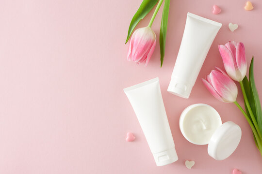 Organic beauty products concept. Top view photo of cosmetic tubes without label open cream jar colorful hearts and pink tulips on pastel pink background with copy space
