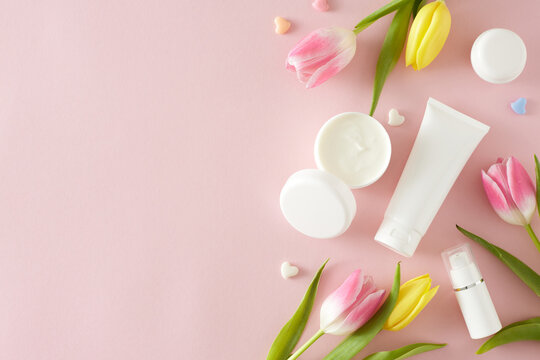 Organic beauty products concept. Top view photo of open cream jar cosmetic tubes colorful hearts and yellow pink tulips on pastel pink background with empty space