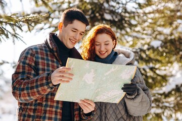 Fototapeta na wymiar A married couple stands in the middle of a snowy forest and studies a map. A smiling red-haired woman and her boyfriend are studying a map to make a route through the forest.