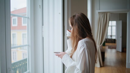 Fototapeta na wymiar Calm woman sipping coffee at flat interior closeup. Girl watching window posing