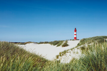 Fototapeta na wymiar Red and white striped tall building of lighthouse against blue clear sky. Sand ground with green grass in foreground.