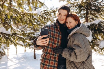 A happy young couple takes a photo together in front of snow-covered trees. Smiling young people decide to take a picture while walking on a day walk in a snow-covered park.