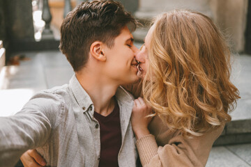 Young cheerful couple is taking a selfie while giving a kiss to each other outdoors.