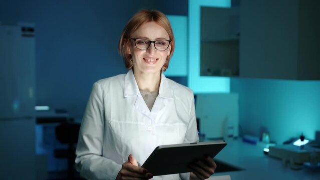 A veterinarian in white lab coat and glasses looking up from a tablet computer and smiling. She is in a laboratory and is wearing glasses. She looks focused. High quality 4k footage 