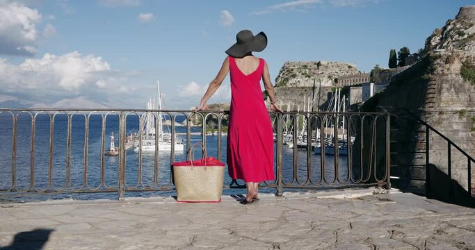 Fashion model woman at in front of Old Fortress at Kerkyra with yachts docked in marina, Corfu Island, Greece, She wear colorful red dress and black hat