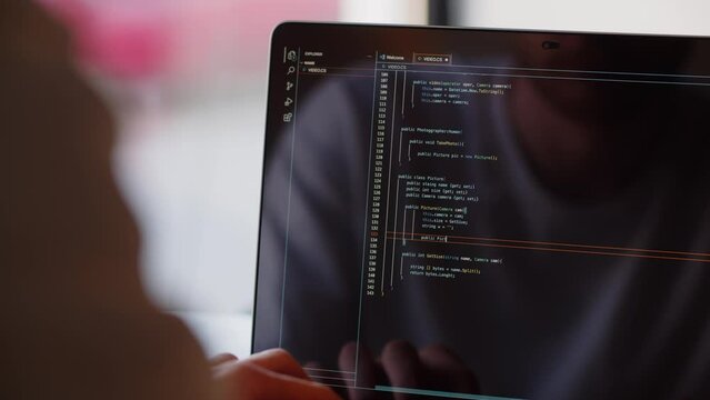 Rear view of a male developer writing code for a website. The programmer works remotely behind his laptop, sitting in a cafe.