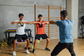 Unrecognizable trainer instructor shows lifting exercise with dumbbell weight in a big gym, concrete wall background. Teenagers and personal coach performing circuit routine training. Horizontal