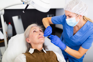 Female cosmetologist in mask and medical gloves making injections of botulotoxin in face of elderly woman.