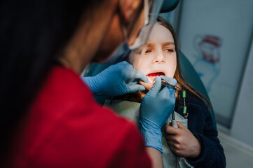 Dentist woman in rubber gloves with tools diagnoses, treats teeth from caries of a girl, a child in a clinic, dentistry office. Photography, medicine.