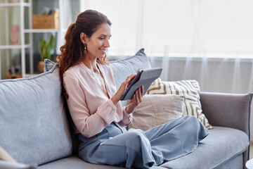 Side view portrait of smiling adult woman using digital tablet while sitting on couch at home and shopping online or using internet