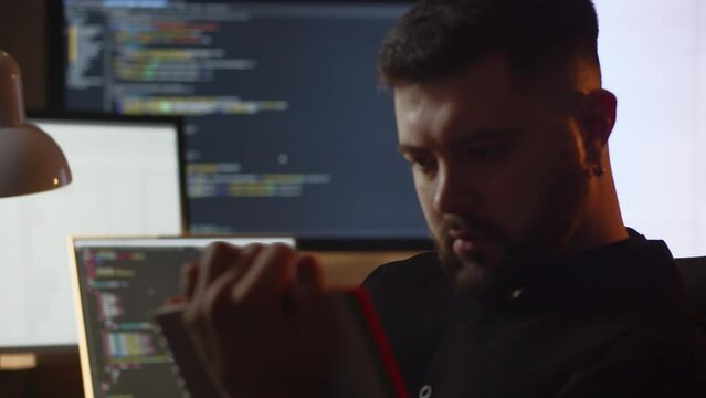 A stylish young man with an earring in his ear sits in front of monitors with program code. Close-up of a male programmer writing something in a notebook.