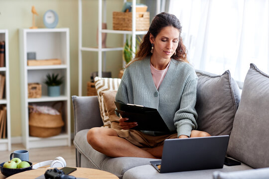 Full Length Portrait Of Young Woman Using Laptop At Home While Sitting On Sofa Holding Clipboard, Copy Space