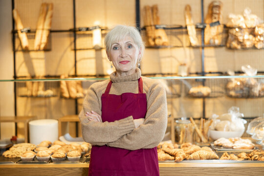 Confident elderly bakery saleswoman in maroon apron posing with arms crossed indoors in front of glass display case with fresh pastries