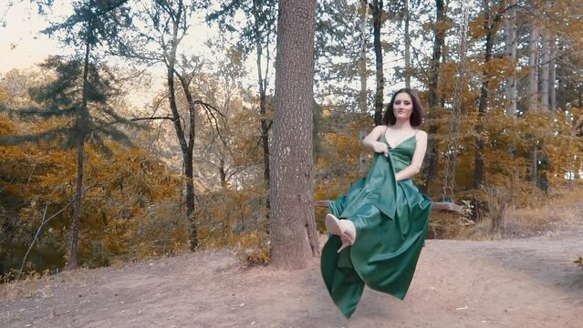 Young beautiful woman sitting on the swing in a luxurious, green silk dress, with a long train and posing in the royal park. Shooting for elegant prom and events dresses. Slow motion