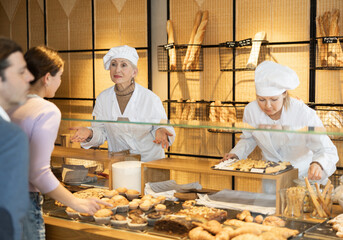 Polite smiling aged female baker in white uniform working behind counter in small private bakery shop, offering customers large assortment of fresh artisanal pastries