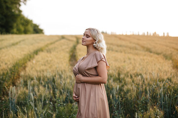 A pregnant blonde woman in a long dress is touching her belly in a wheat field at sunset.	