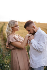 A man with a beard in a white shirt is kissing hands of his pregnant wife in a dress in the middle of the field.