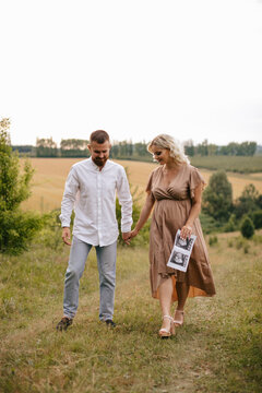 A pregnant woman in a long dress is holding ultrasound baby image and walking with husband in a field at sunset. Future parents.	