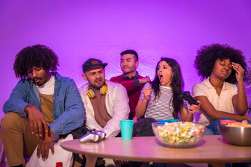 Adult party. Young people sitting on the sofa playing video games with popcorn. Joystick or controller in hand, purple led, woman celebrating race victory