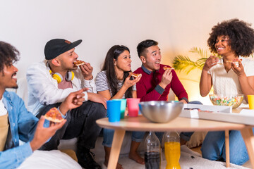 Group of multiethnic friends on a sofa eating pizza and drinking soda at a home party, eating pizza and telling jokes