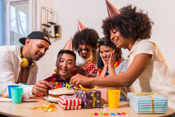 Multi-ethnic group of friends at a birthday party on the sofa at home with a cake and gifts,...